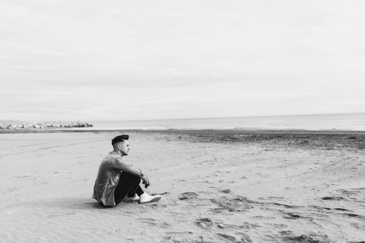 A Grayscale Photo Of A Man Sitting On The Beach