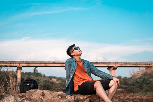 Free Full body of young male tourist with black backpack in casual wear looking at blue sky while lounging on stones by bridge in sunlight Stock Photo