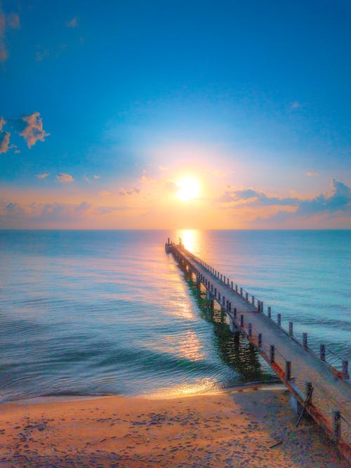 Long old narrow pier above footprints on sandy beach and colorful blue sea under vivid blue sky at sunset