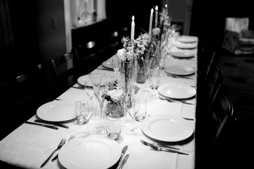 Black and white cutlery near round plates and glasses by decorations on long narrow arranged holiday table for guests in dark room