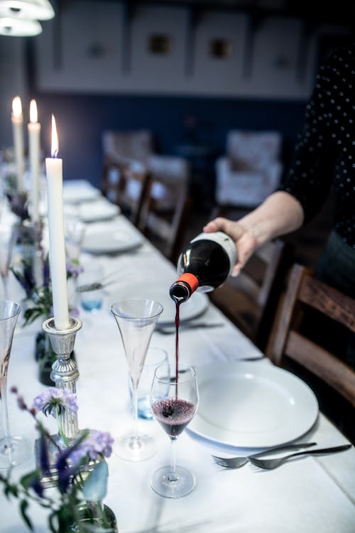 Crop person in black shirt pouring dark red wine in transparent glass on white table decorated with plants and flaming candles