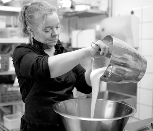 Black and white of focused adult female chef in black wear with hair tied up pouring white liquid eatable mass in big glistening metallic dish