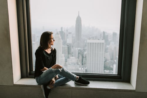 Free Woman in Black Jacket and Blue Denim Jeans Sitting on Window with the View on Empire State Building Stock Photo