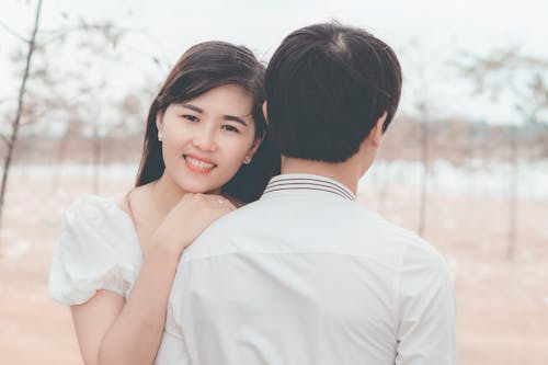 Smiling Asian girlfriend hugging unrecognizable boyfriend while standing in village in summer day