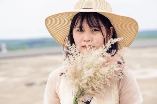 Tranquil Asian female in straw hat and with bouquet of dried grass standing on beach on blurred background of sea and looking at camera