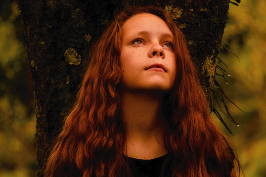 Portrait of a young woman with ginger hair and freckles looking up thoughtfully against a tree.