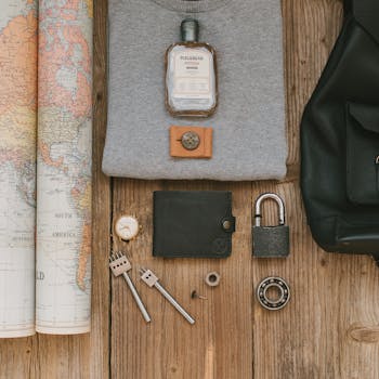 Flat lay of travel items including a map, wallet, watch, padlock, and fragrance on wooden surface.