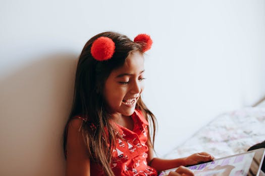 Happy girl in red shirt and pom-pom headband using a tablet indoors with a smile.