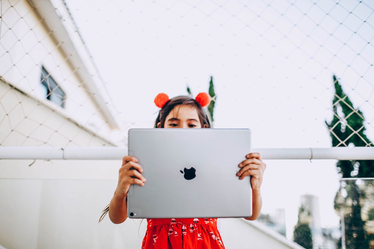 a young girl stands outdoors holding a large tablet wearing a bright r