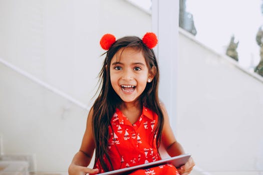 A happy young girl in a festive red dress with pom-poms, laughing and holding a tablet indoors.