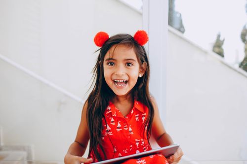 Free A happy young girl in a festive red dress with pom-poms, laughing and holding a tablet indoors. Stock Photo