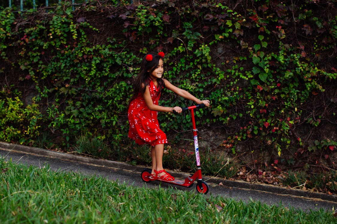 Side view of content child in dress riding kick scooter along road in park in summer 