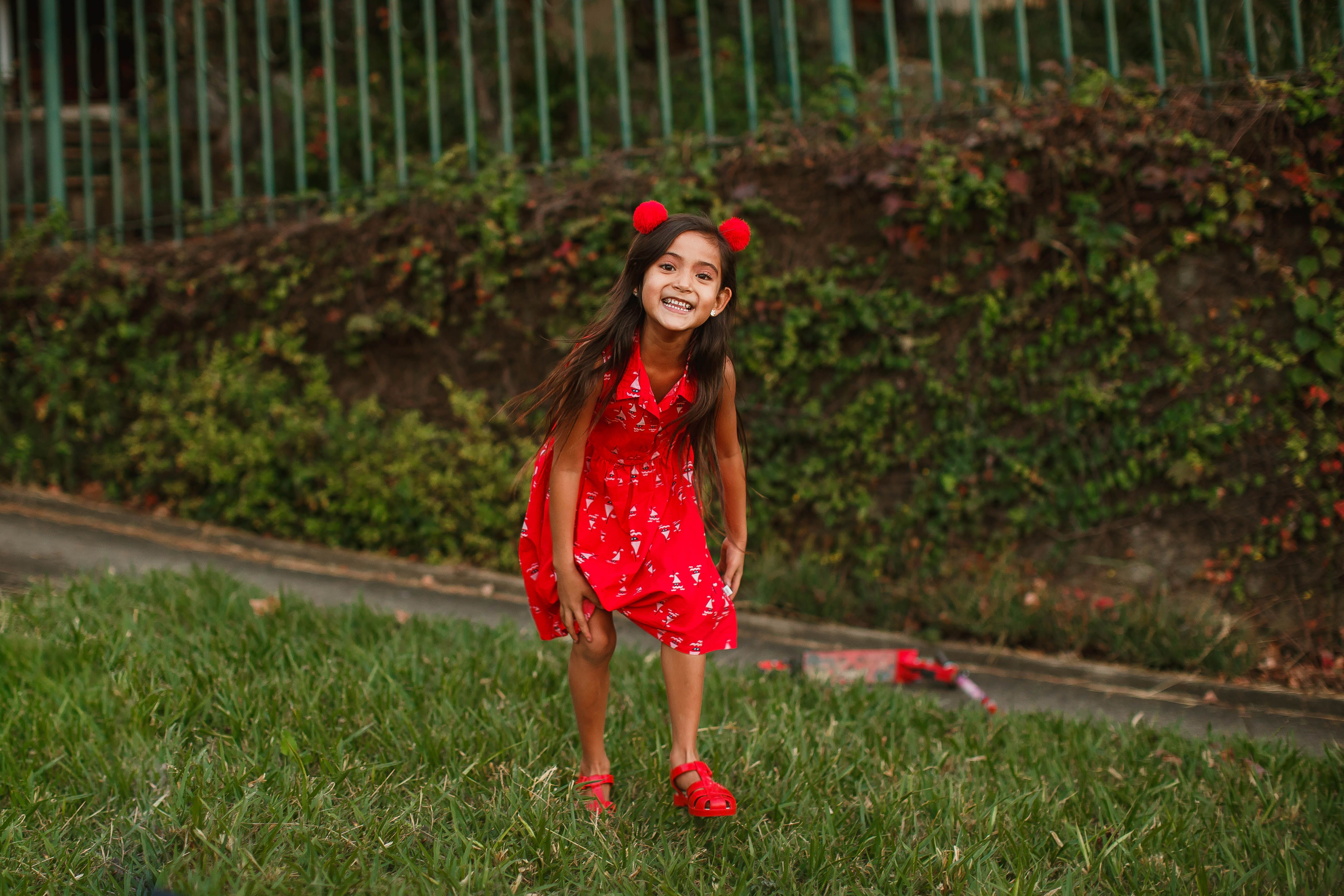 Adorable girl in red summer dress standing on green lawn and cheerfully looking at camera