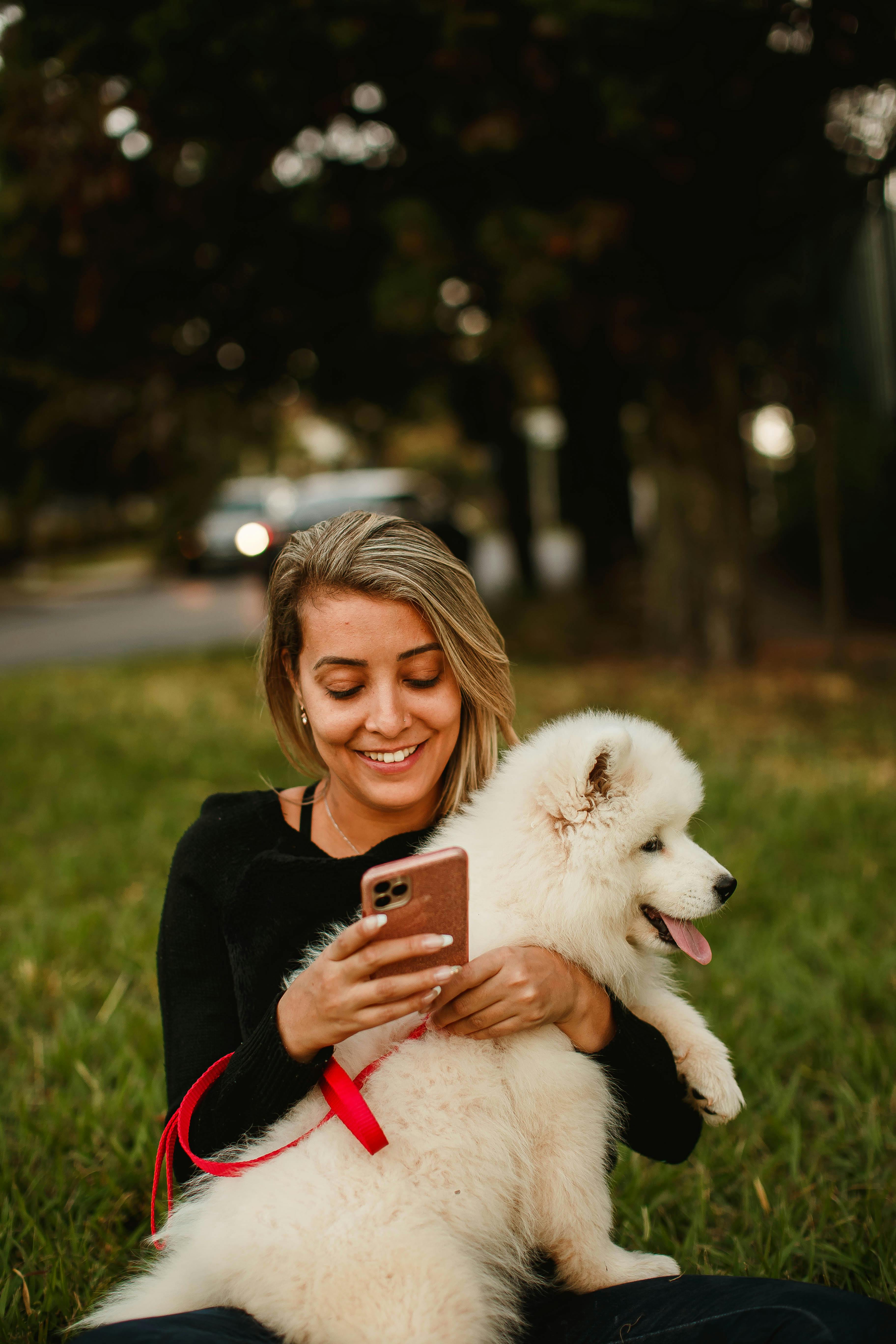 Cheerful female owner sitting on lawn with charming fluffy dog and browsing cellphone during stroll in park