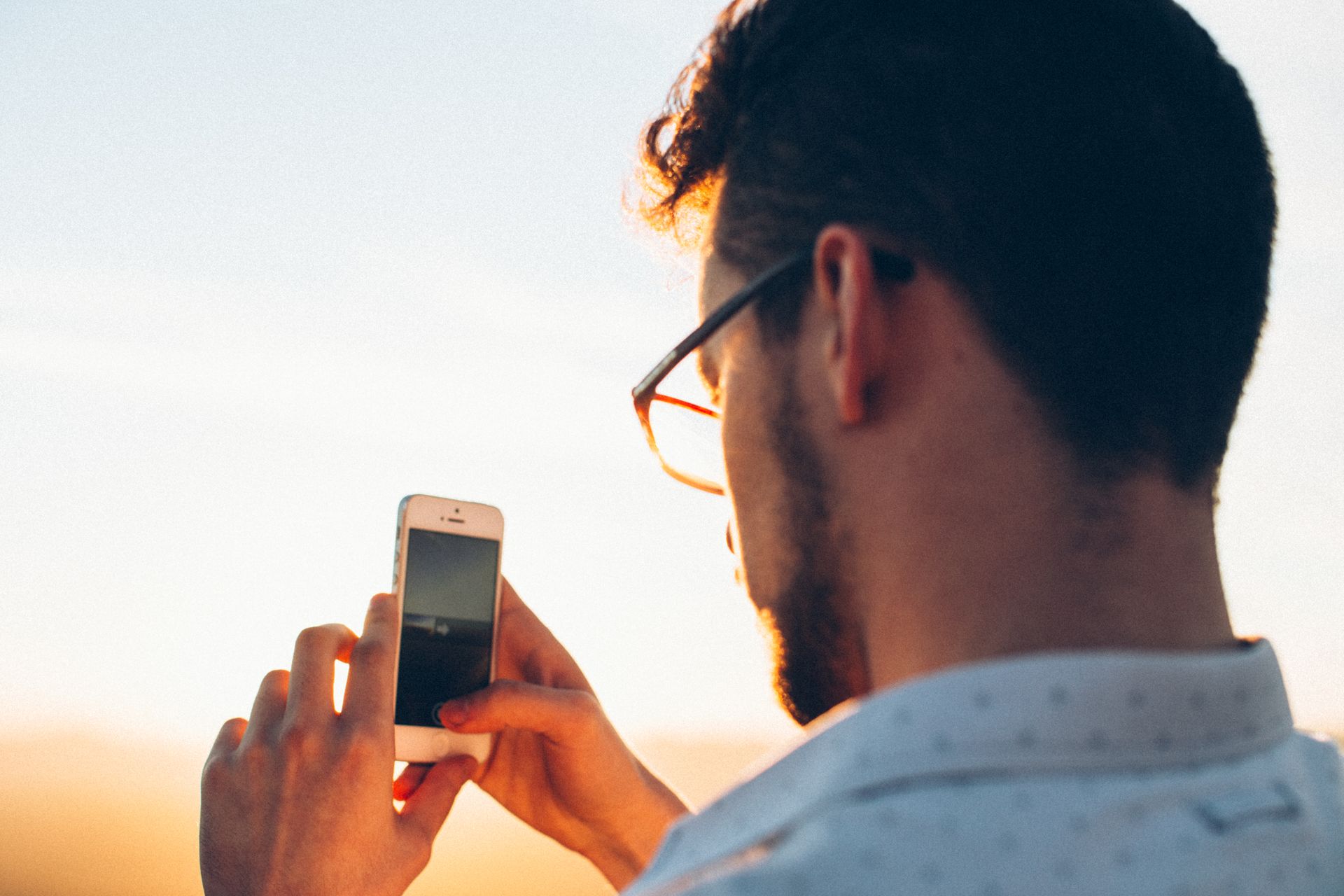 Back view of serious young bearded male in casual wear and glasses taking photo of scenic morning landscape on digital mobile phone