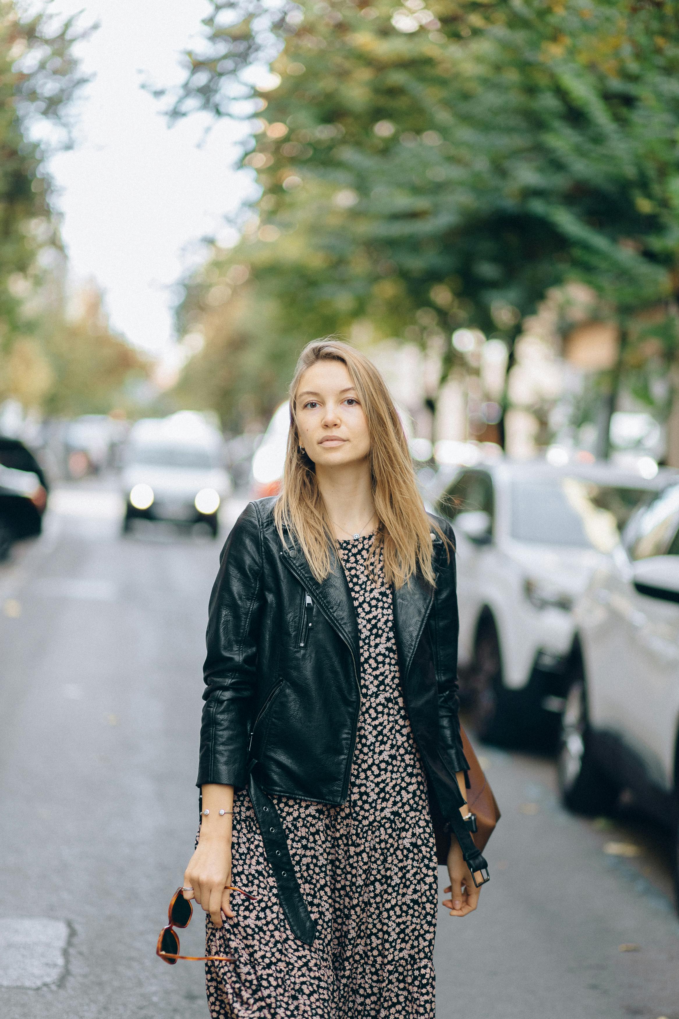 Young woman in floral dress and leather jacket strolling down a city street.