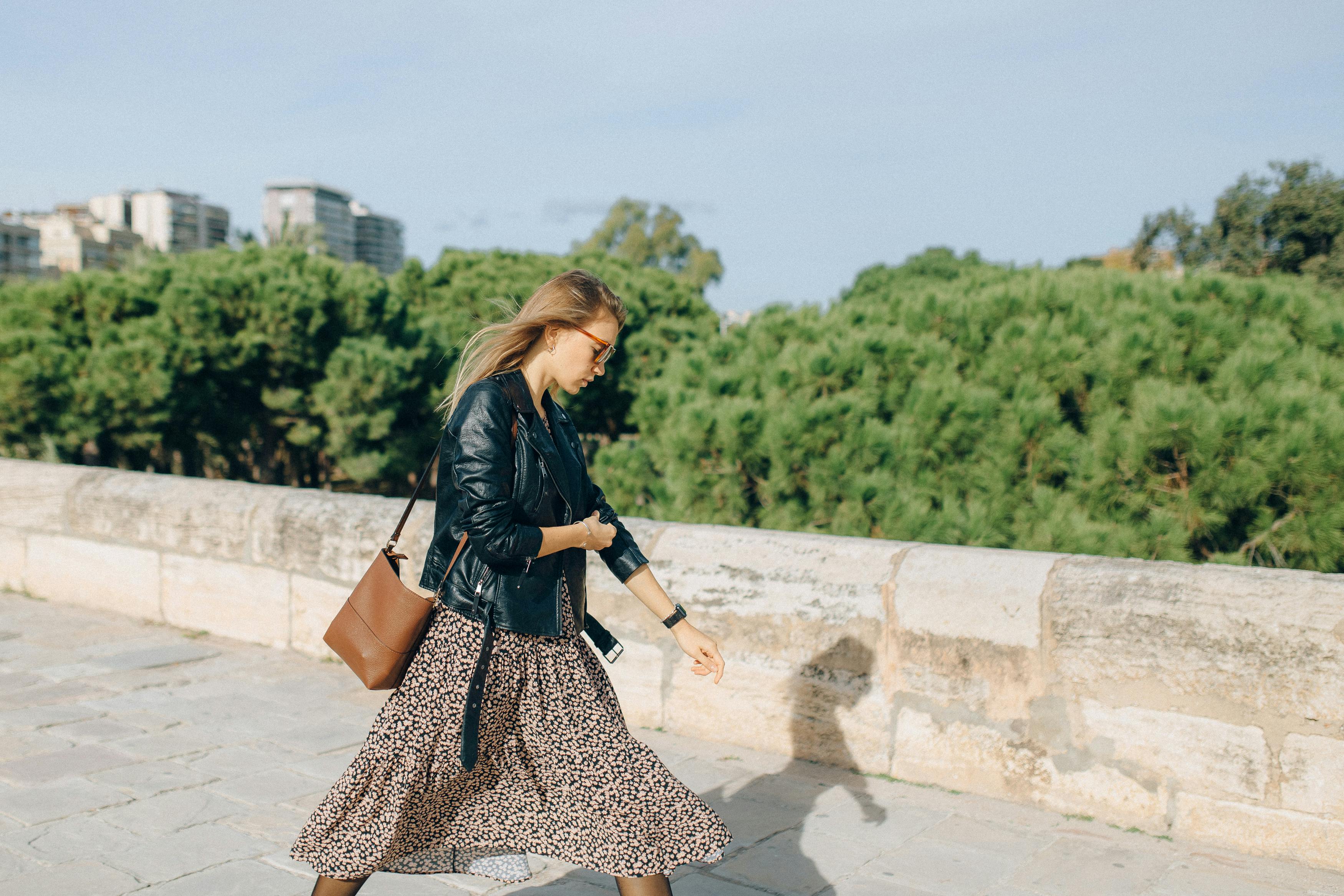 Woman in sunglasses, a black leather jacket and floral skirt walking along a stone bridge with trees in the background
