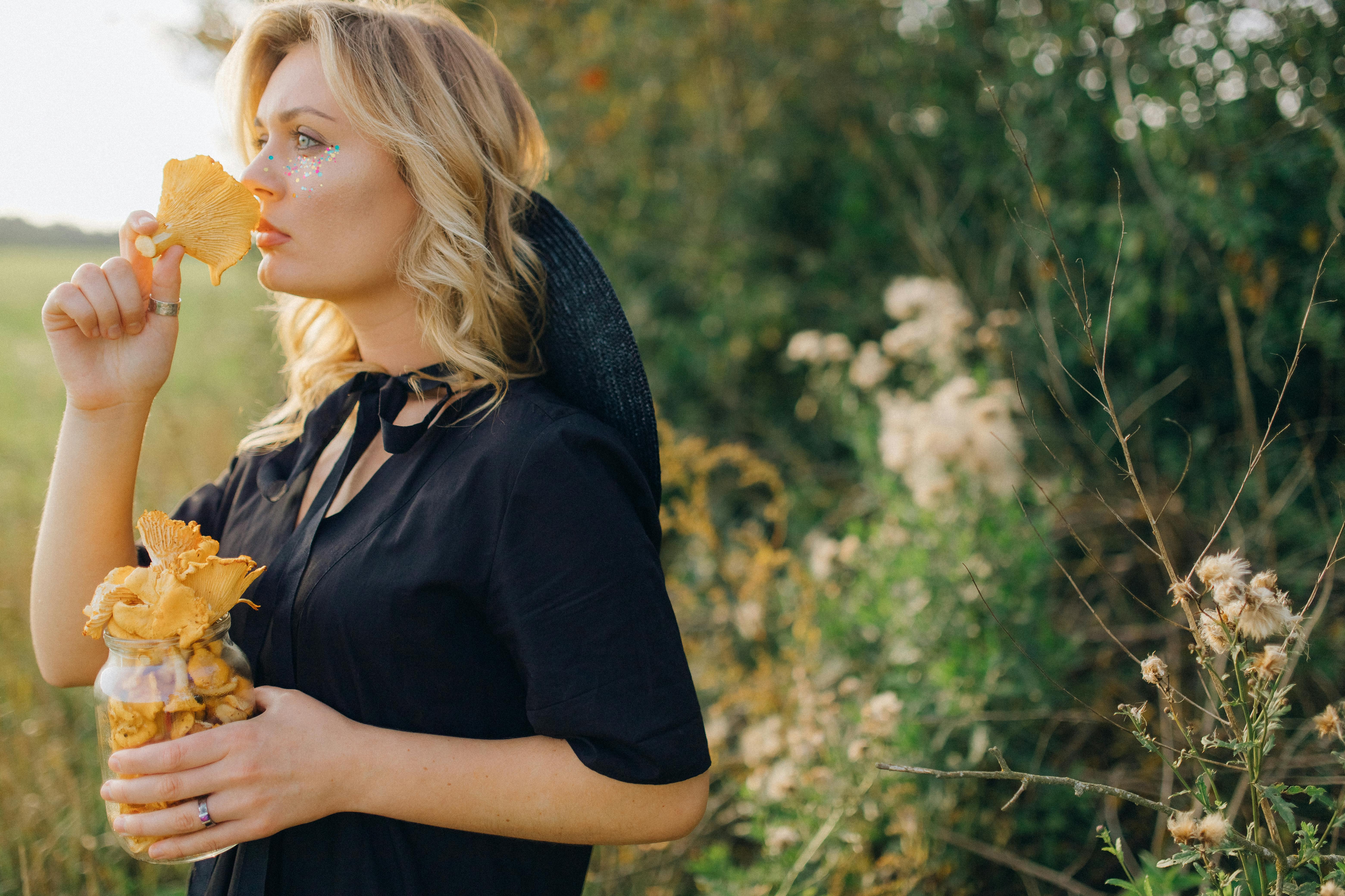 Blonde woman with glitter makeup holding chanterelle mushrooms in a sunny field.