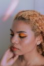 Young mindful ethnic female with colorful makeup standing with closed eyes while touching face near wall