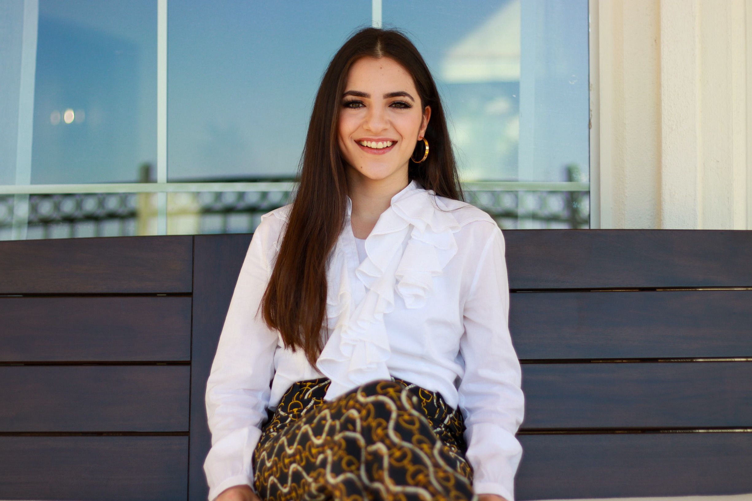 Elegant young woman with long hair smiling while posing outdoors in Austin, wearing a white blouse and patterned skirt.