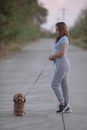 Side view of smiling ethnic female in sportswear holding small fluffy dog with tongue out by leash on walkway
