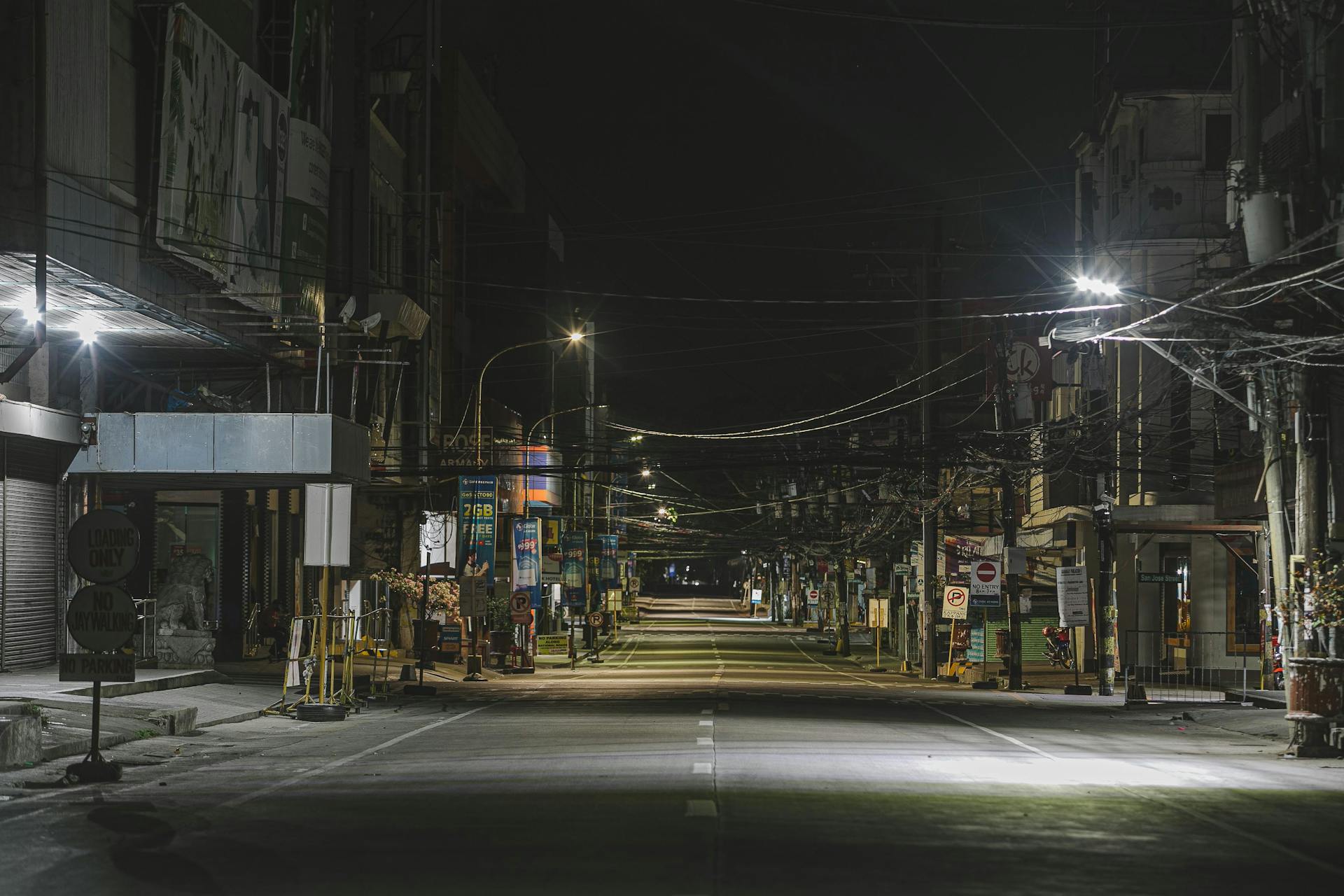 Straight asphalt road between city houses with shining lamp posts and wires at night