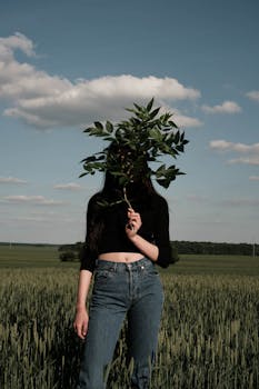 在乡村多云天空下，一位身穿休闲装的匿名女性站在草地上，用绿叶遮住脸