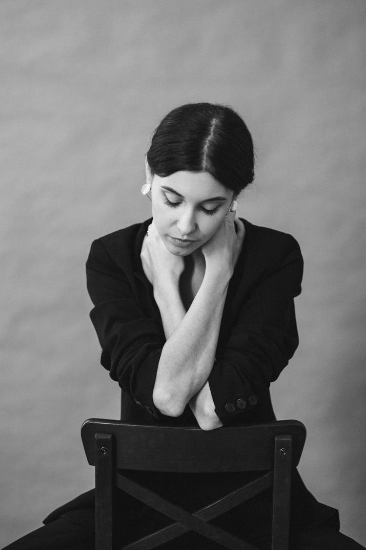 A Grayscale Photo Of Woman In Black Blazer Sitting On The Chair With Her Hands On Her Neck