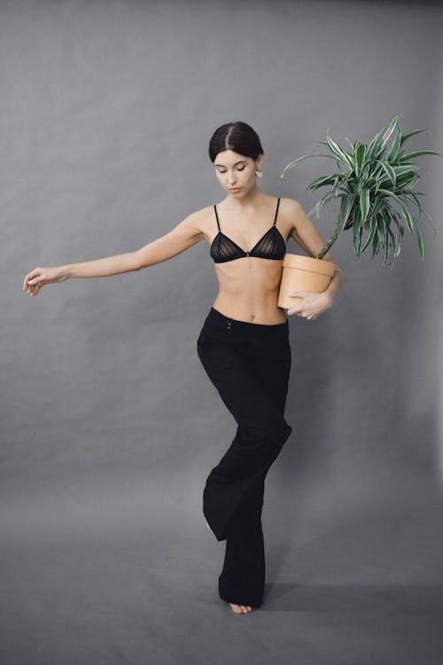 Woman in Black Brassiere and Black Pants Holding a Pot of Green Plant while Looking Down