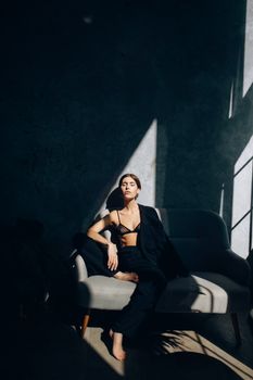 An elegant woman sitting in a dark room, wearing a black ensemble and relaxing on a sofa, sunlit shadows enhance the mood.