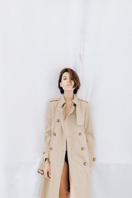 Striking fashion portrait of a woman in a beige trench coat against a white background, showcasing modern elegance.