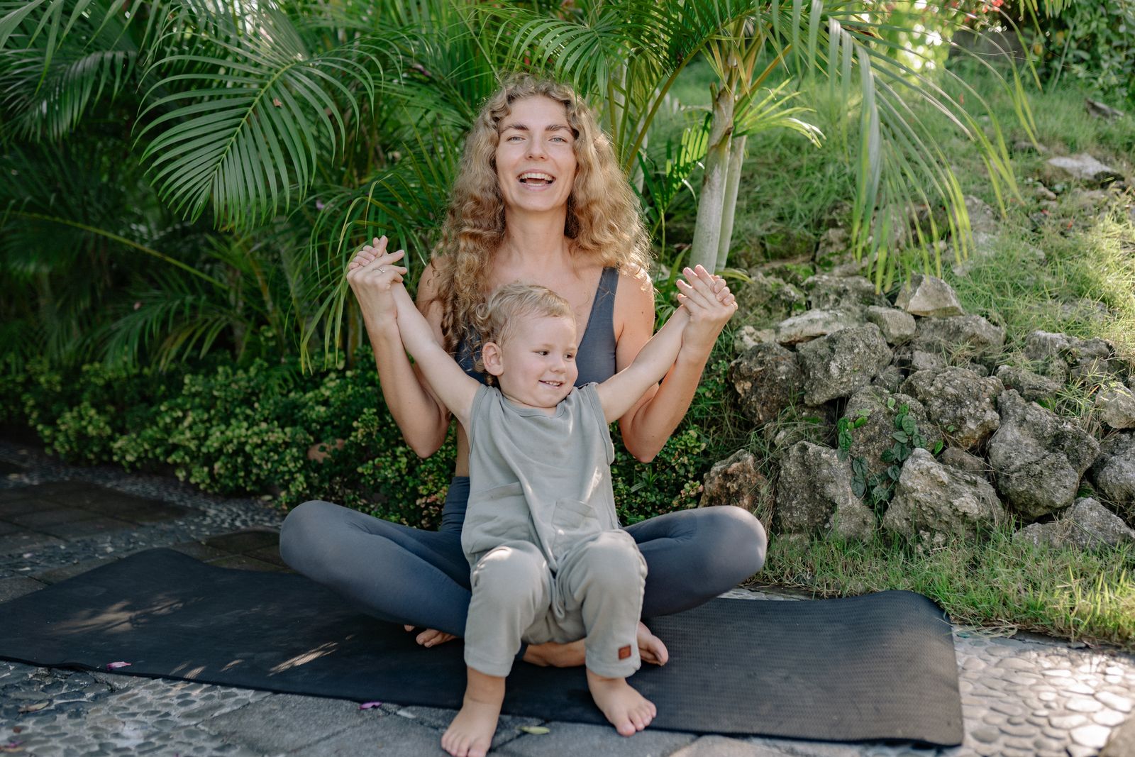 Joyful mother practicing postnatal yoga outdoors with her baby on a mat surrounded by greenery
