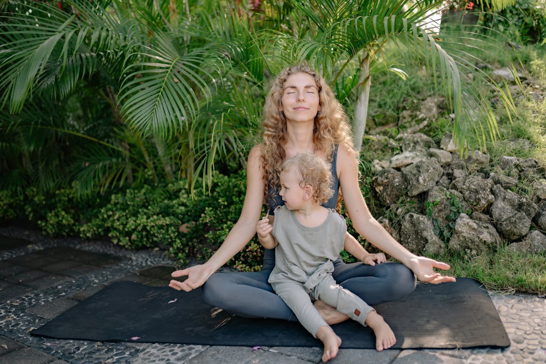 Free Full body happy relaxed mother sitting with eyes closed in lotus pose with cute boy on laps while practicing yoga in tropical green park Stock Photo