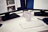 White Ceramic Mug Between Apple Magic Keyboard and Two Flat Screen Computer Monitors