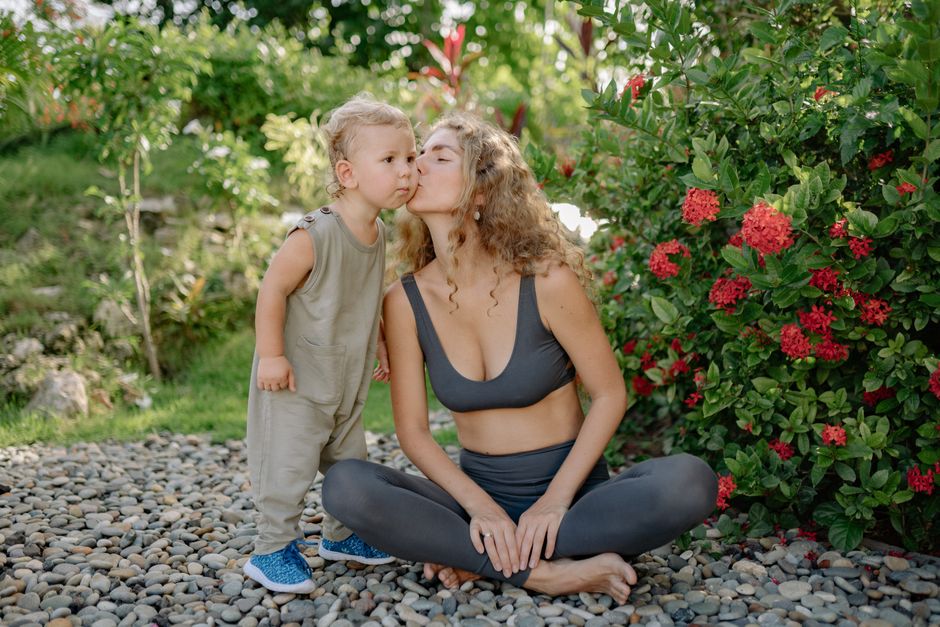 Full length positive sportswoman in gray leggings and sports bra sitting in lotus pose and kissing cute little son cheek after practicing yoga in green kush garden