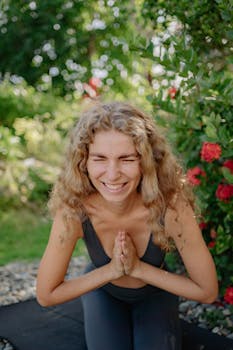 A joyful woman practicing yoga outdoors with hands in prayer position, surrounded by greenery.