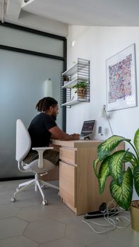 African American man working remotely at home office desk with a laptop, surrounded by plants.