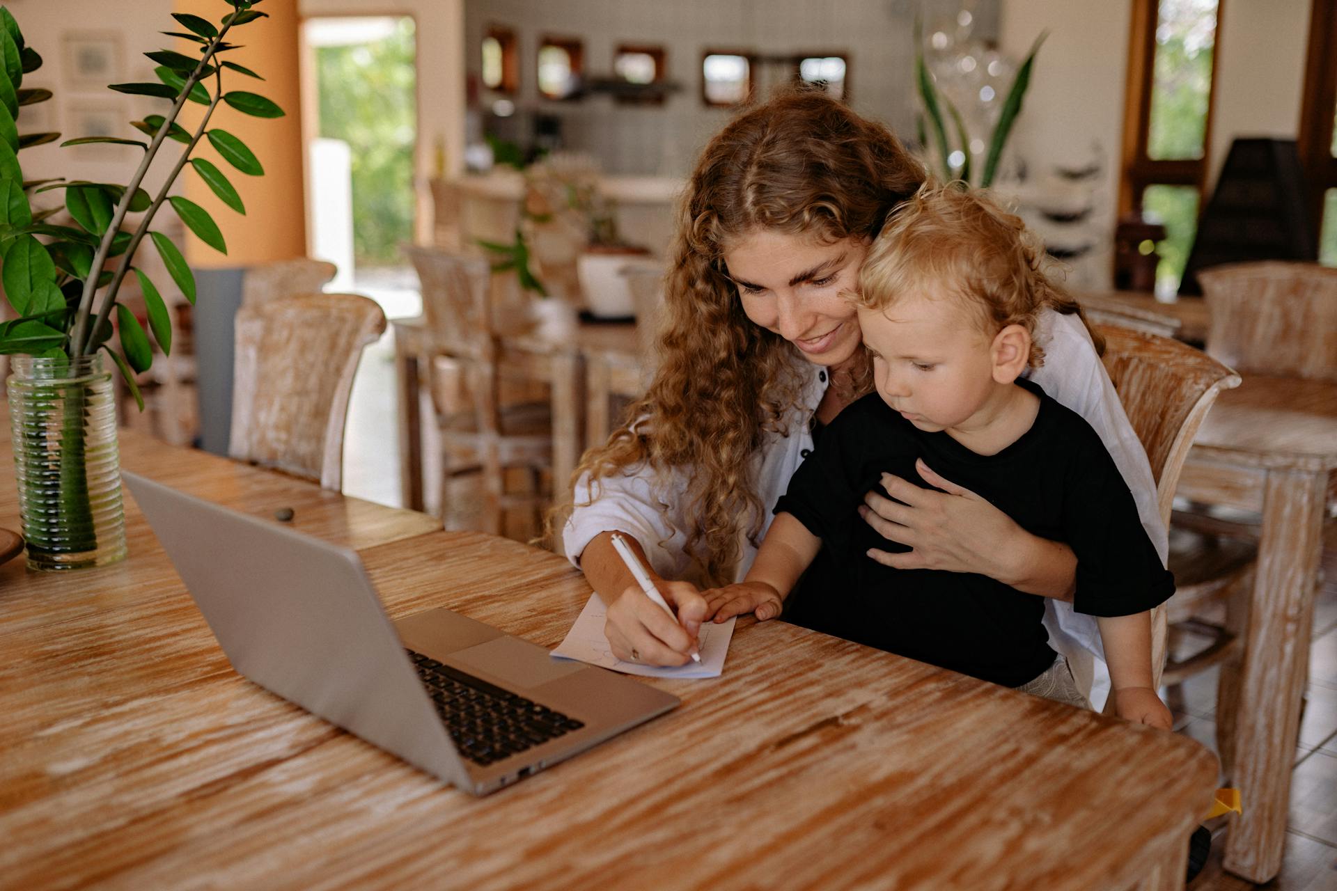 A mother assists her young son in writing while using a laptop at a wooden table.