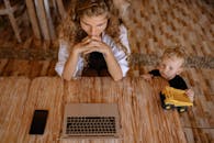 一名妇女正坐在木桌前用笔记本电脑工作，旁边有个孩子在玩玩具卡车