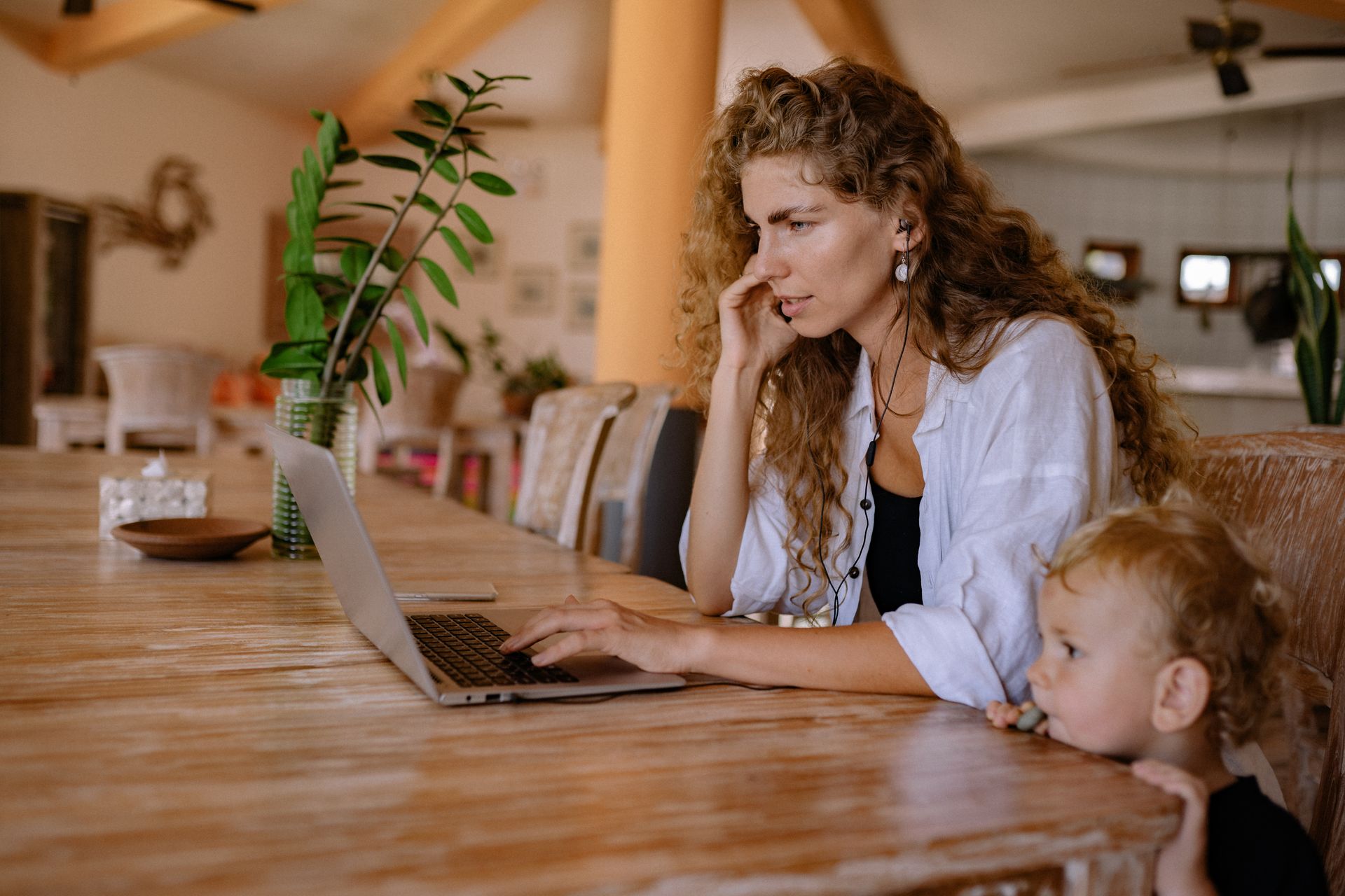 Woman working on a laptop with her child beside her at a wooden table in a cozy home environment.