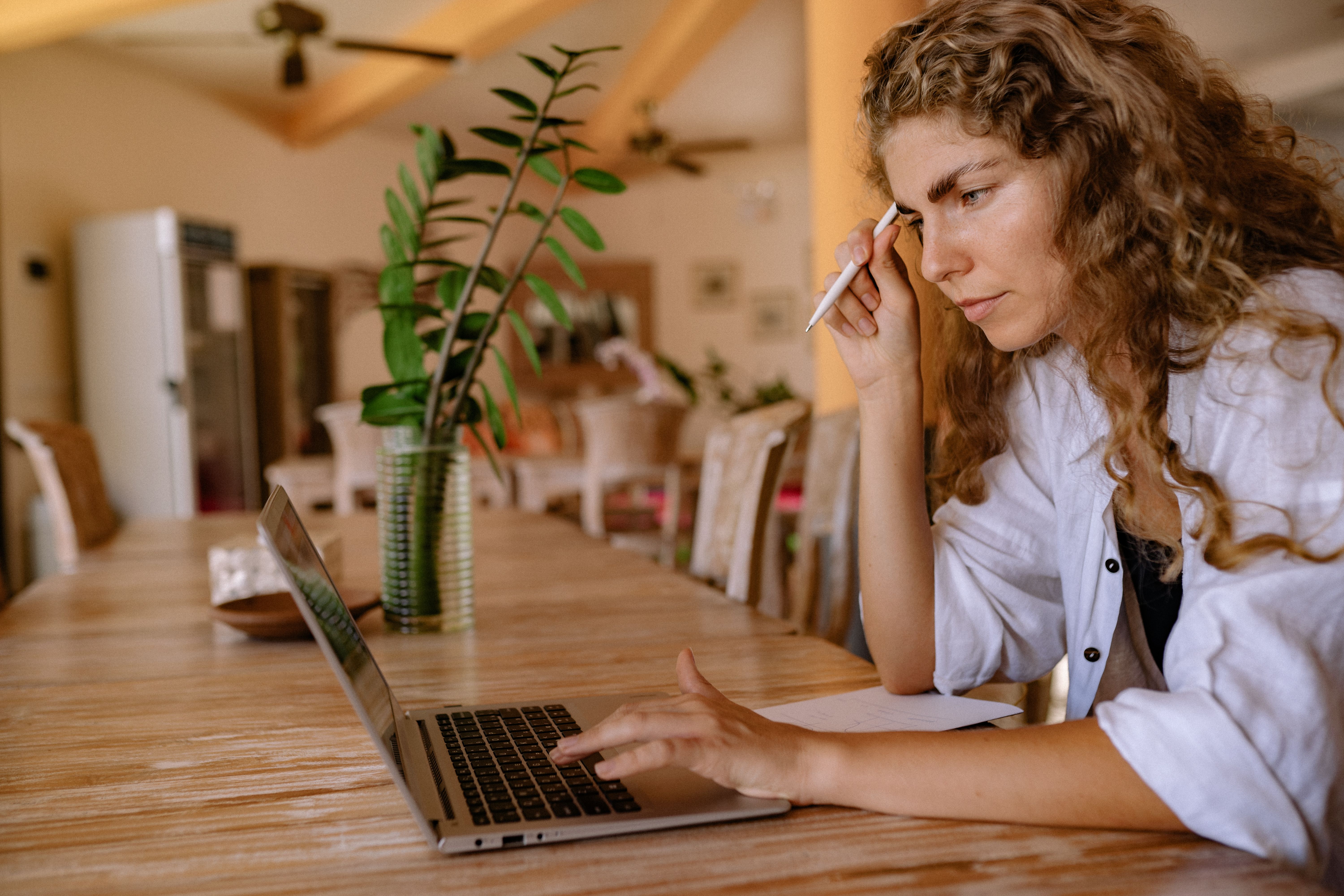 Focused woman with curly hair using laptop at home table, multitasking and working remotely.