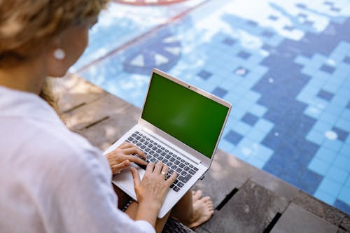 Free From above of crop unrecognizable barefoot female remote worker surfing internet on netbook with chromakey screen while sitting near swimming pool in sunlight Stock Photo