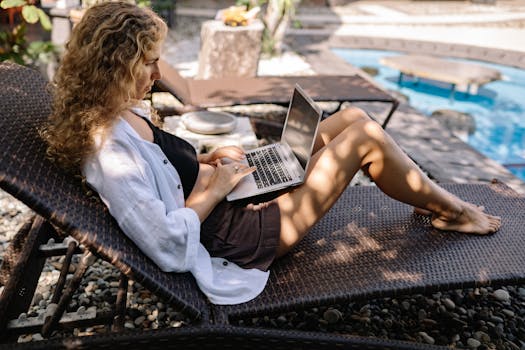 Woman with curly hair working on her laptop outdoors, relaxing on a sunlounger by the pool.