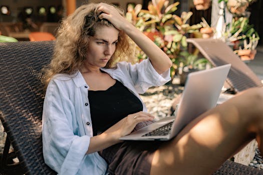 Focused woman with curly hair working on a laptop outdoors in a sunlounger, embracing a digital nomad lifestyle.
