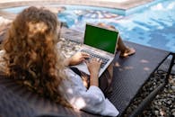 从上图看，一名匿名女性自由职业者躺在游泳池边的日光浴床上，正用便携式电脑打字，阳光下可见透明的水