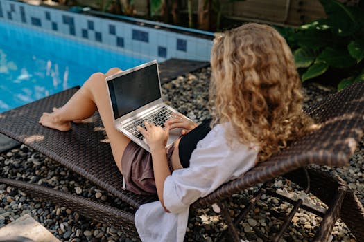 A woman with curly hair works on a laptop while lounging by the pool in a relaxed outdoor setting.