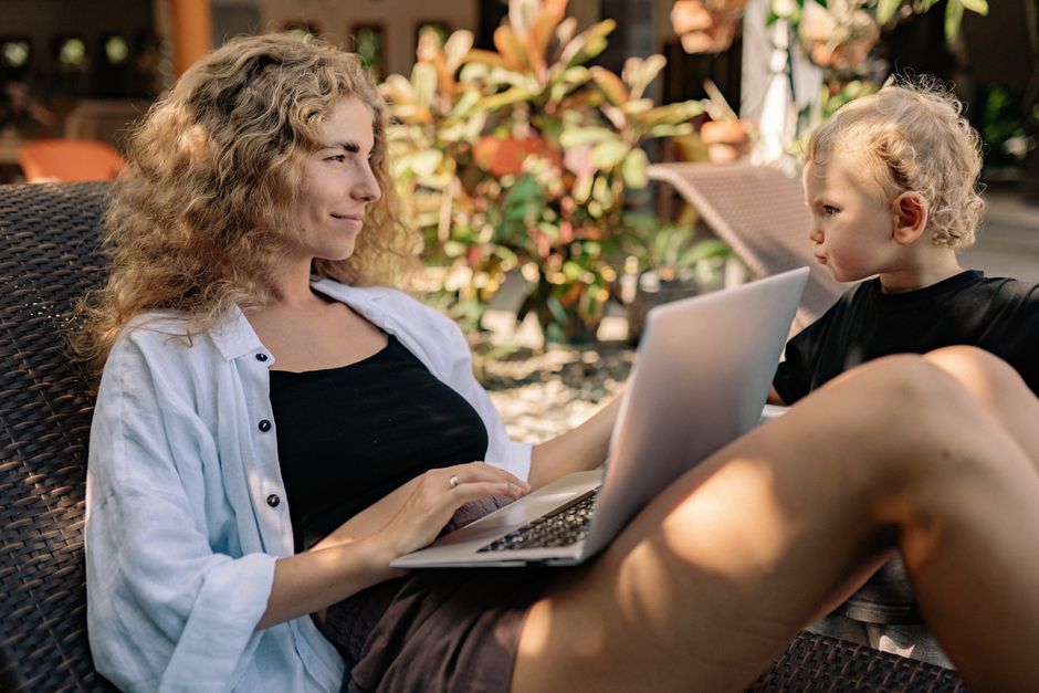What Dermatologists Say About Baby Sunscreen Ingredients and SPF A woman working on a laptop outdoors with a child beside her on a sunlounger.