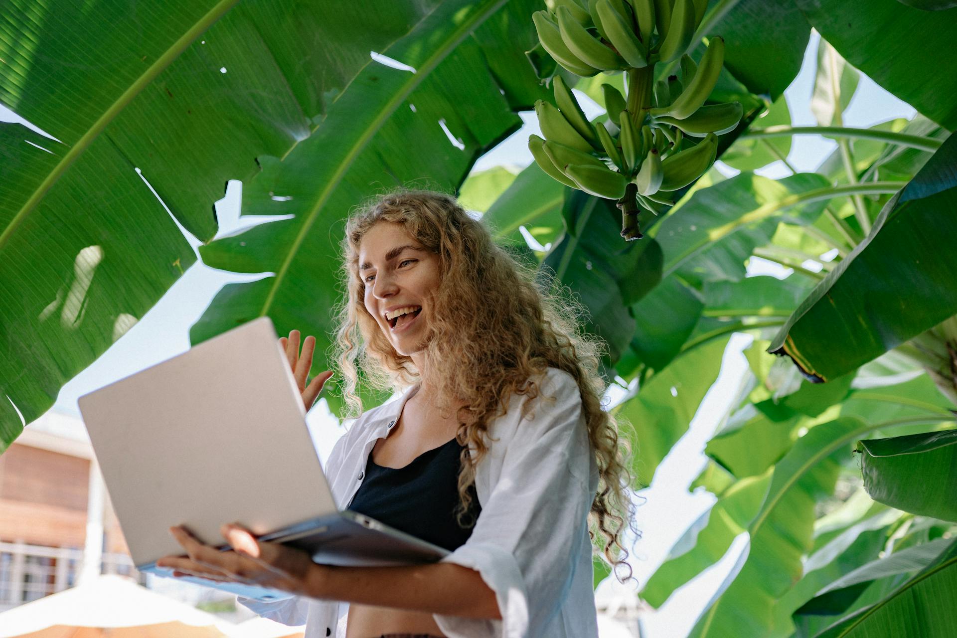 A joyful woman waves during a video call on a laptop in a lush tropical environment.