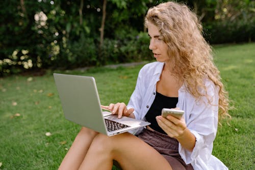 Free Thoughtful female freelancer with smartphone surfing internet on laptop in park Stock Photo