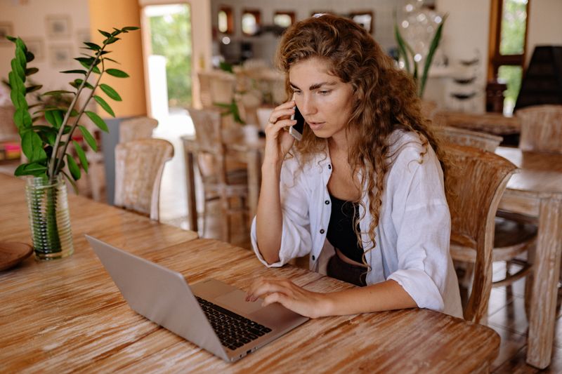 A woman in a bright room using a laptop and phone, blending work with home comfort